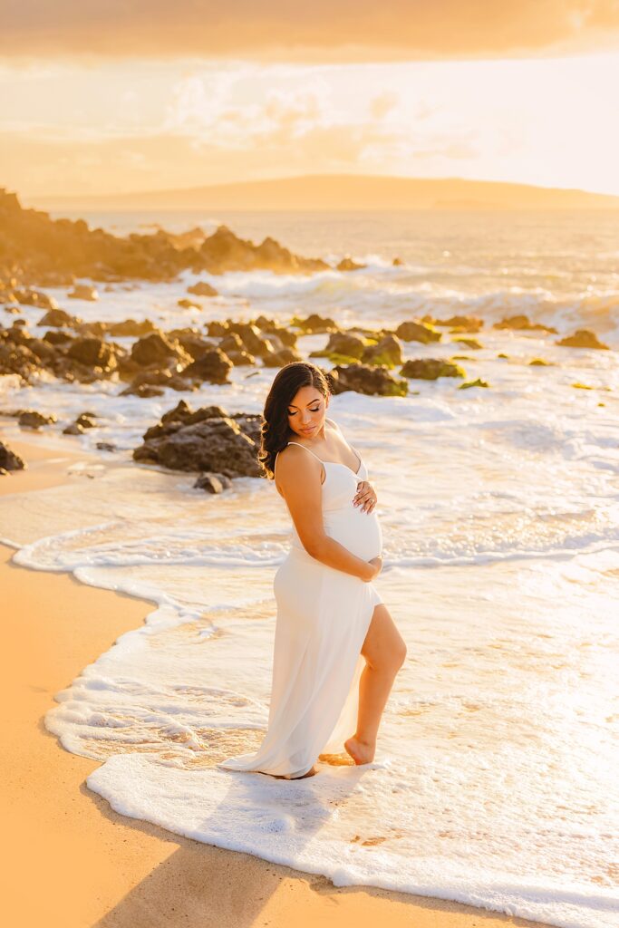 A woman wearing a white maternity dress while standing in the water for beach maternity photos in Hawaii.