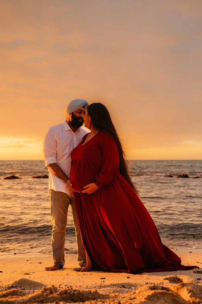A woman wearing a beautiful red maternity dress standing with her male partner on the beach together during a pregnancy photoshoot with love + water photography on maui.