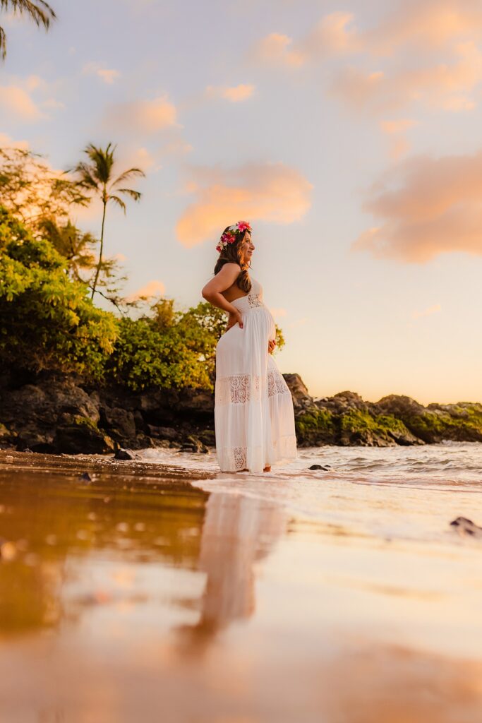 A pregnant woman wearing a white maternity dress and flower crown posing for beach maternity photos in Hawaii.