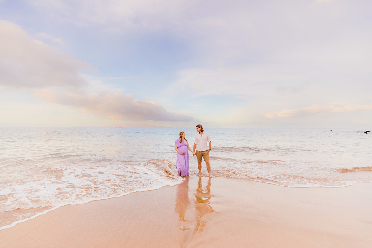 A pregnant woman wearing a light purple maternity photoshoot dress holding hands with her partner while standing in the water for Maui beach maternity photos.