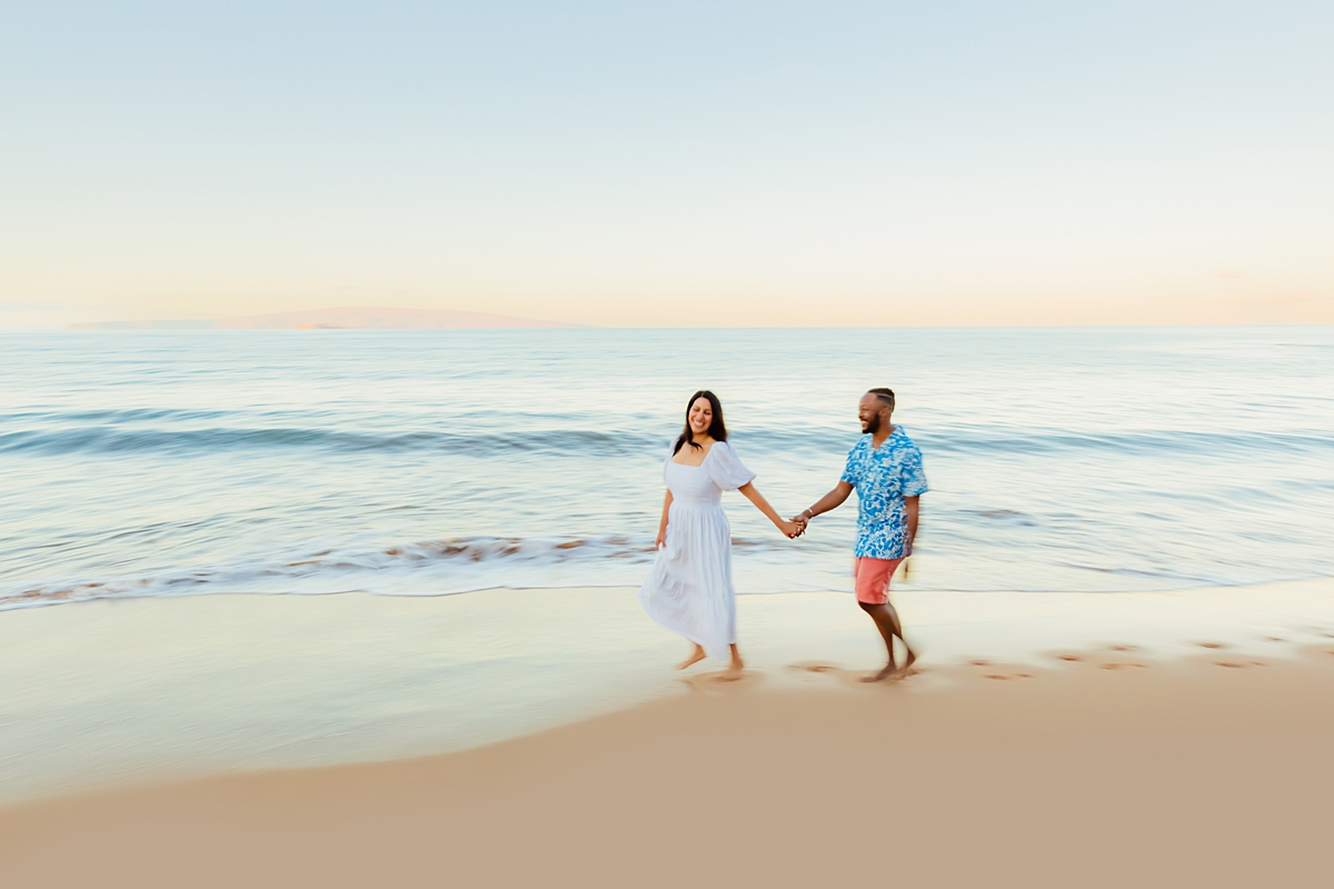 A pregnant woman holding hands with a man walking on the beach for maternity photos.