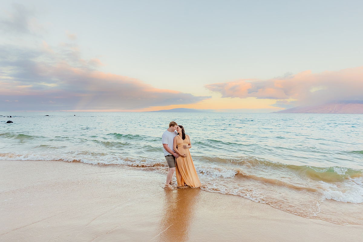 A couple standing together on the shore, the woman wearing a peach-colored maternity dress, during maternity photos on Maui.