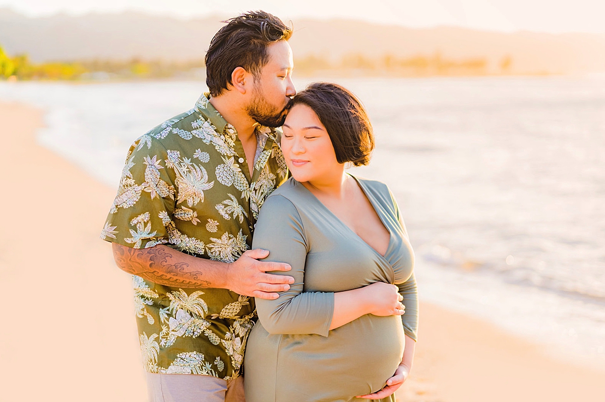 A woman wearing a sage green maternity photoshoot dress while holding her baby bump, and her partner standing behind her and holding her on the beach.