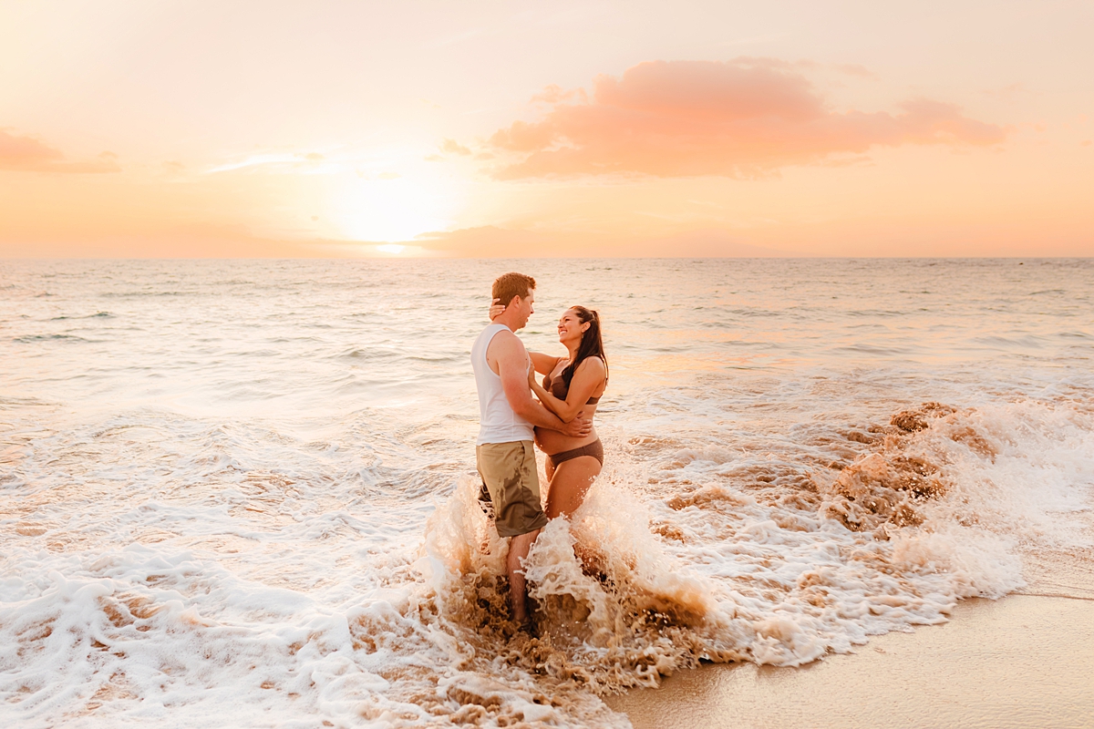 A pregnant couple standing in the water together, the woman wearing a maternity bathing suit, for a maternity photoshoot with love and water photography on maui.