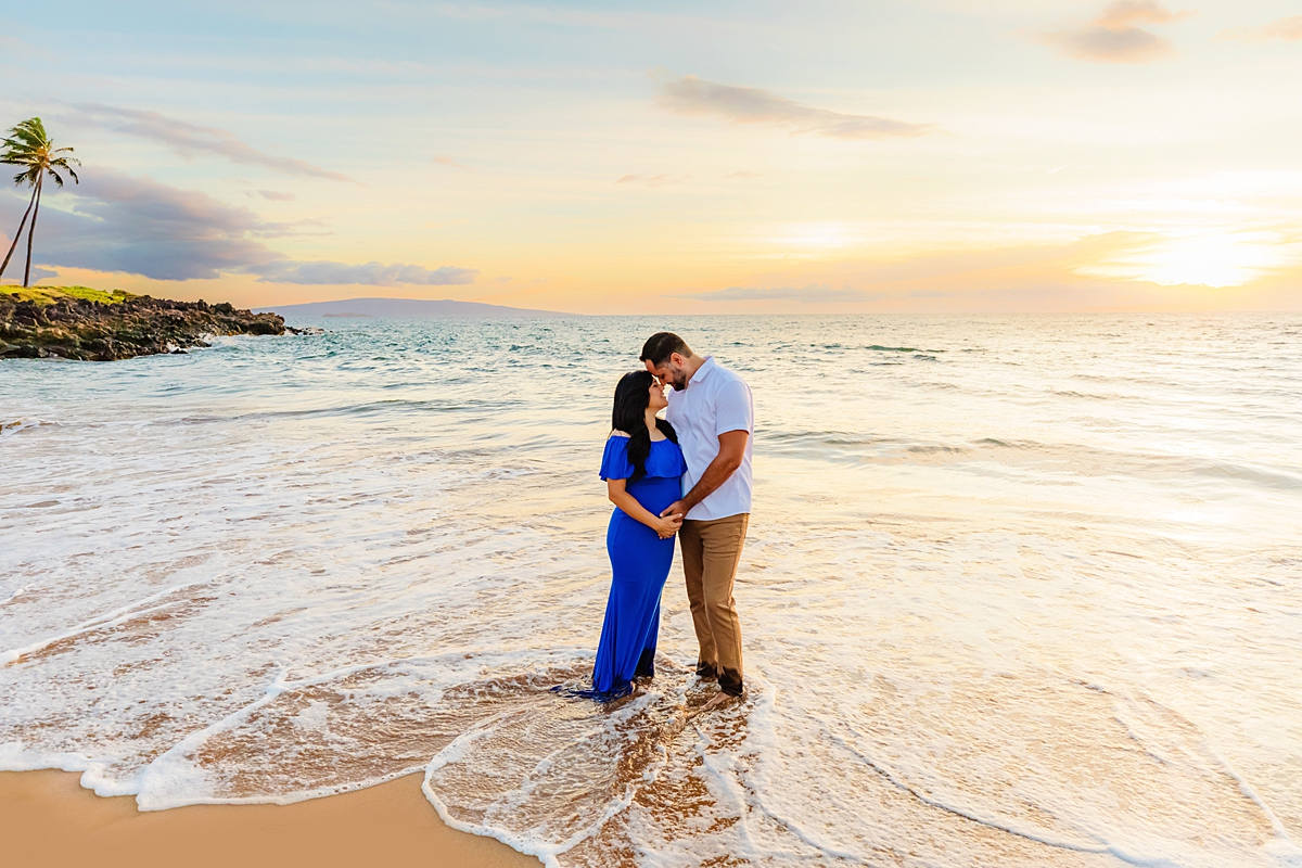 A pregnant couple standing on the beach for maternity photos on Maui, the woman wearing a beautiful blue maternity photoshoot dress.
