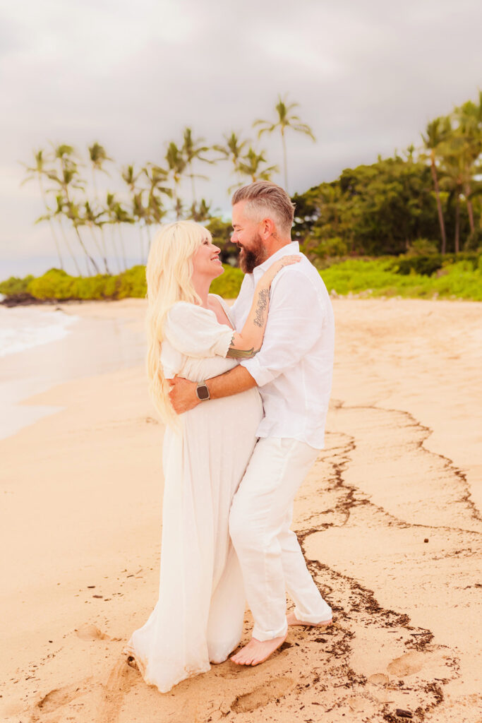 A beautiful blonde woman in a white dress smiles at her husband in a white linen outfit for maternity photos on maui.