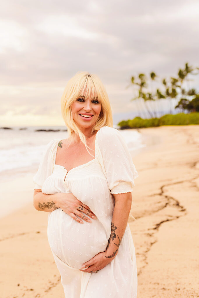 A beautiful pregnant woman with blonde hair in a white dress holds her baby bumb during a maternity photoshoot on the beach on a babymoon on Maui.