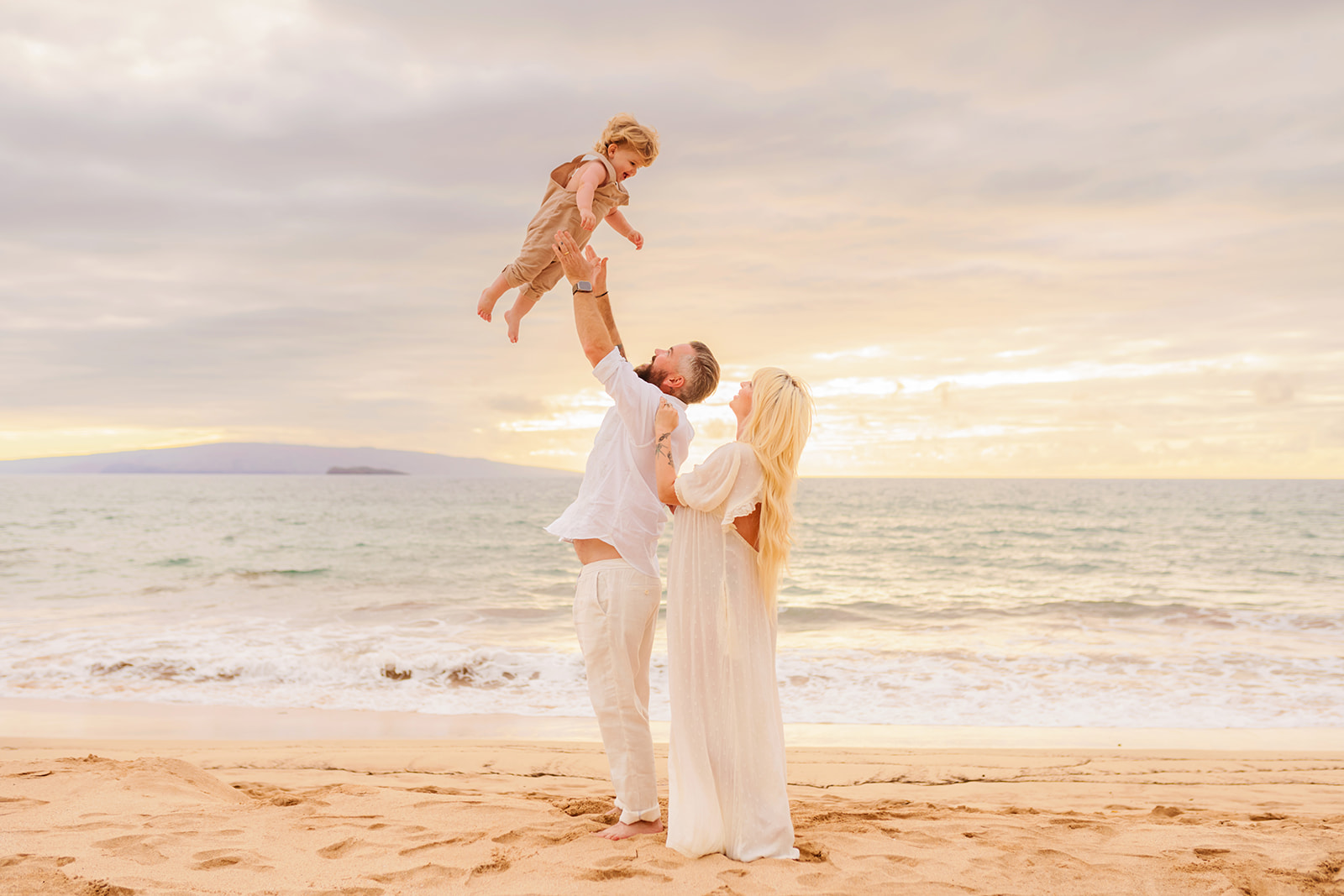 A father throws his toddler in the air while his pregnant wife watches during a maternity photoshoot on the beach on Maui.