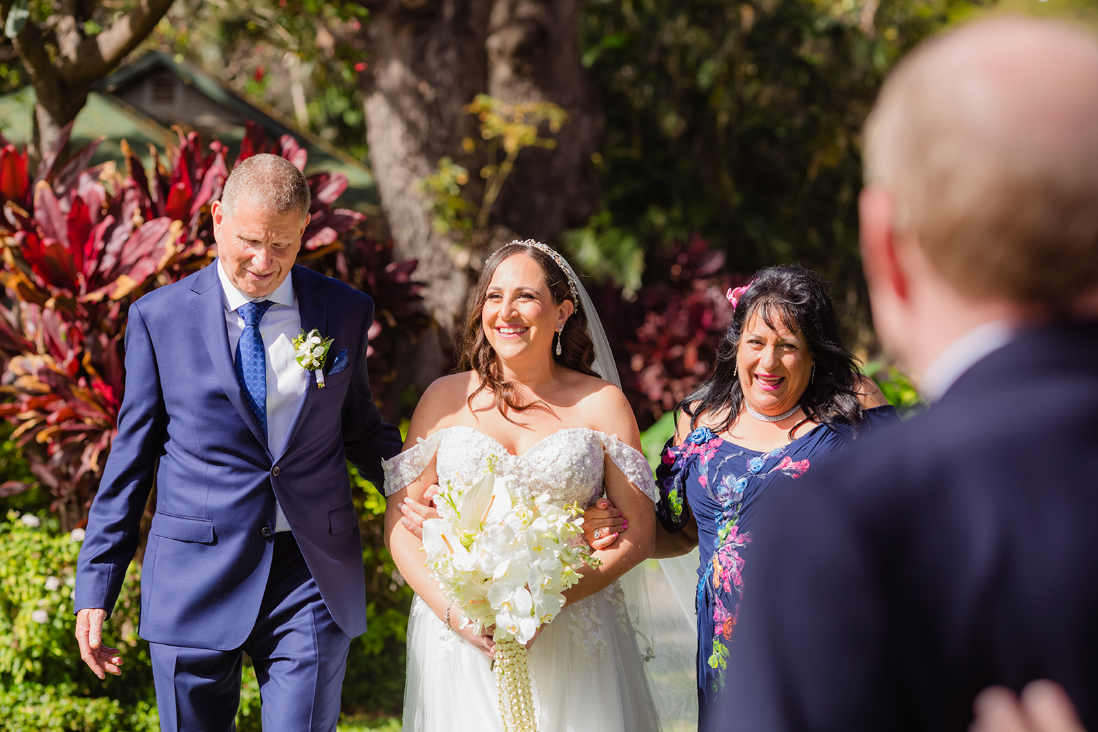 A smiling bride walks arm-in-arm with her mother and father while walking down the aisle for her wedding at Olowalu Plantation House on maui, hawaii.