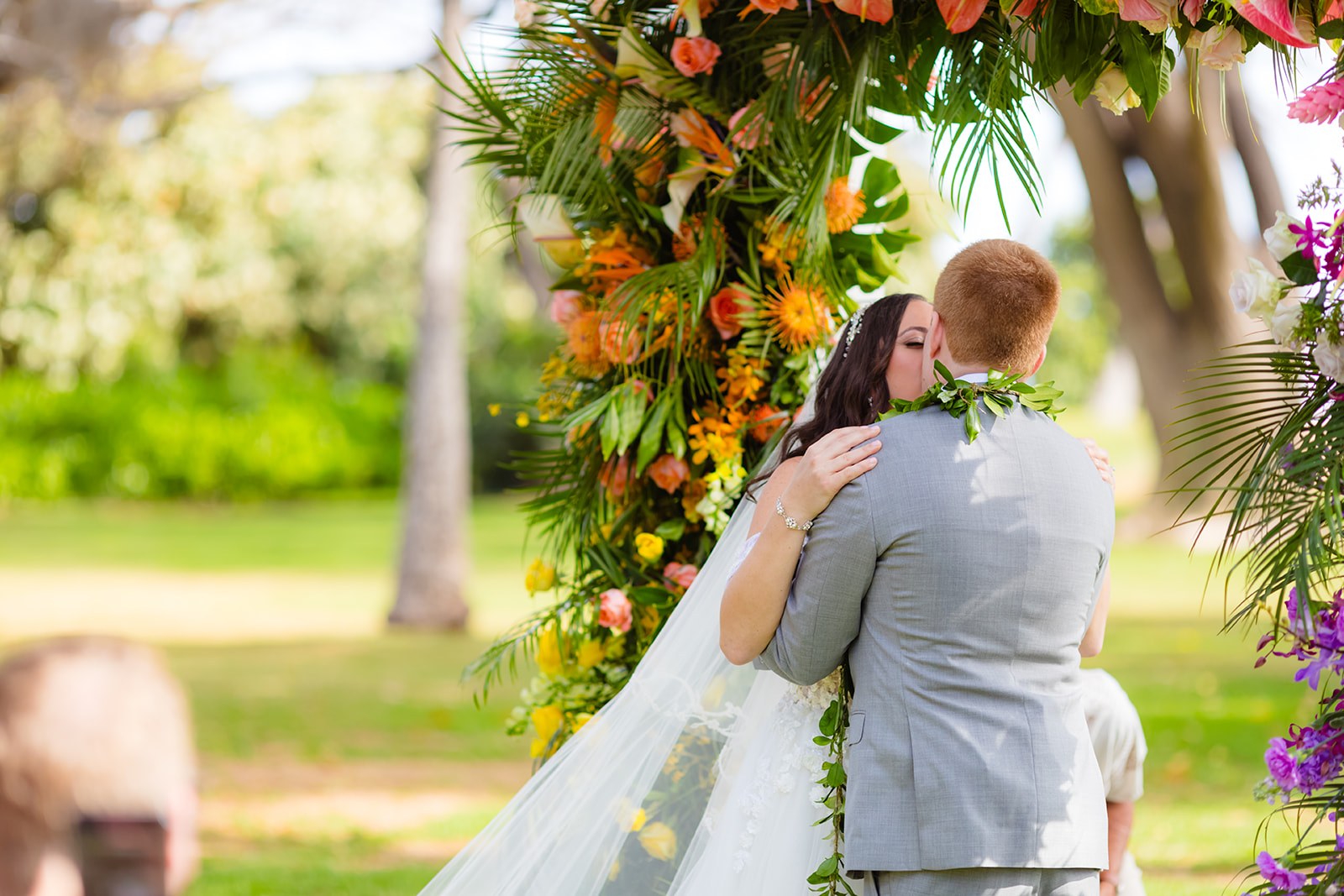 A bride and groom kiss under an arbor of tropical flowers and greenery during a beautiful maui wedding.