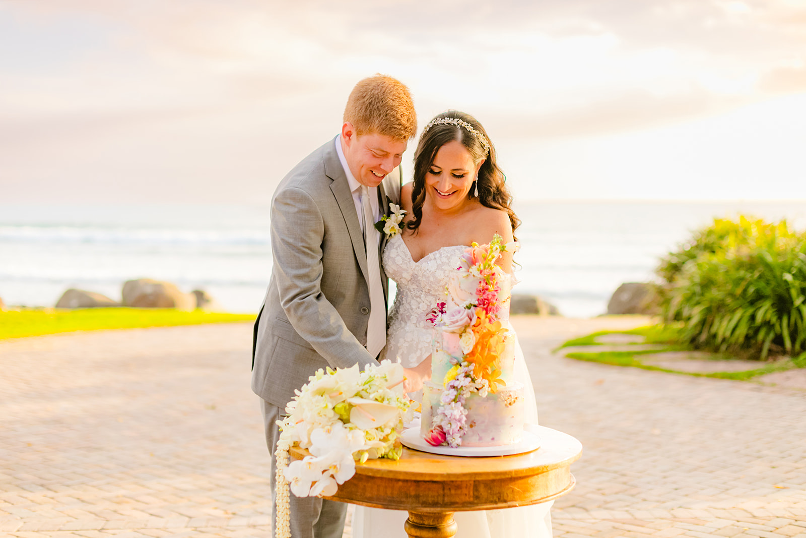 A happy bride and groom slice their wedding cake together in front of a beautiful backdrop of the ocean during a Maui wedding captured by love + water photography.