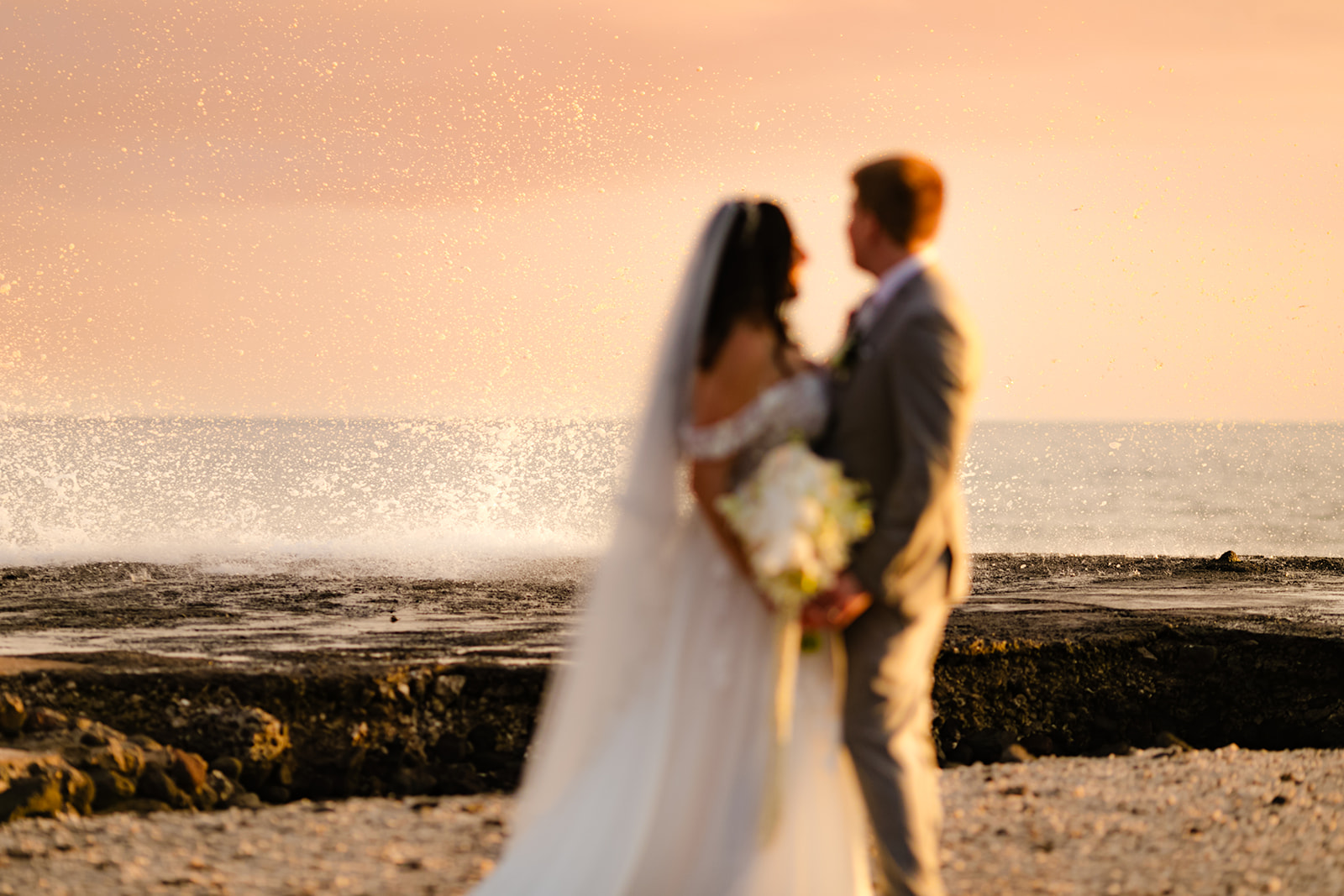 A bride and groom stand in front of the ocean as the water splashes behind them during beautiful wedding photos on maui near Olowalu Plantation House.