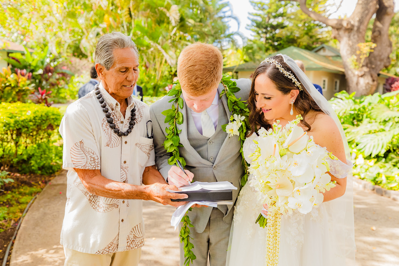 A bride in a beautiful while veil and white bouquet stands beside her groom in a gray tuxedo and their wedding officiant as they sign their wedding documents at Olowalu Plantation House on maui.