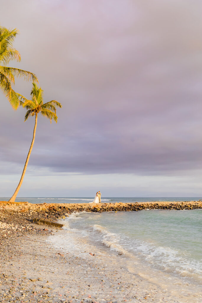 In the distance, a bride and groom stand together on a small peninsula jutting into the ocean during beach wedding portraits on maui, hawaii.