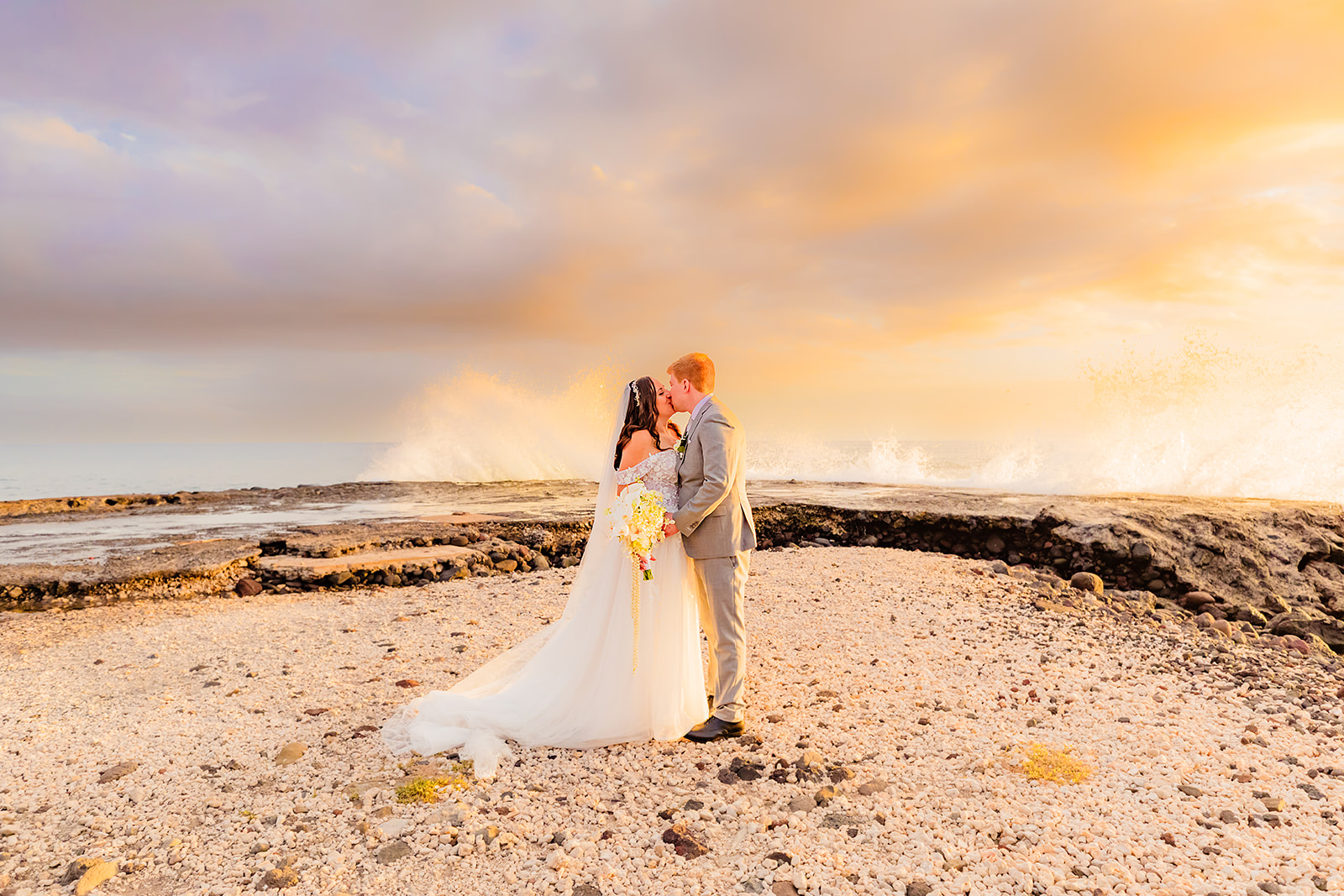 A bride and groom kiss while standing on the beach in front of the ocean during a beautiful sunset as maui wedding photographer love + water photography capture the moment. 