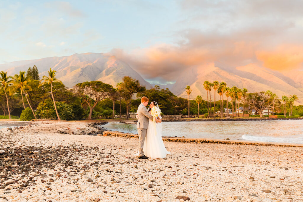 A married couple stands in front of beautiful mountains, palm trees, and the ocean during beach wedding portraits on Maui with love + water photography.