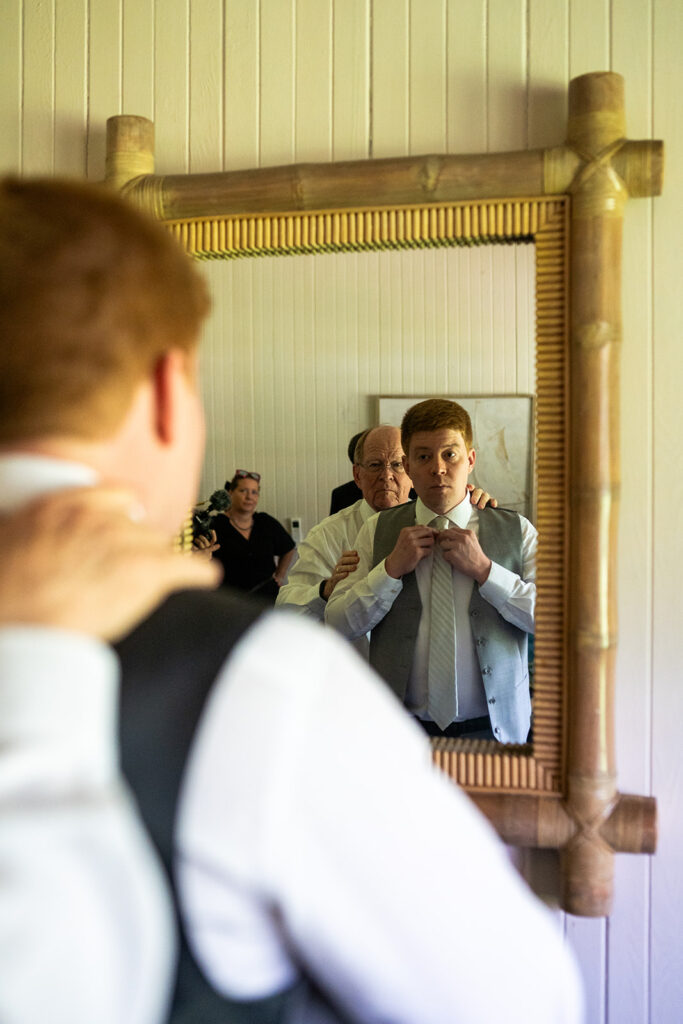 A groom looks in the mirror while his father helps him get ready for his wedding on maui at Olowalu Plantation House.