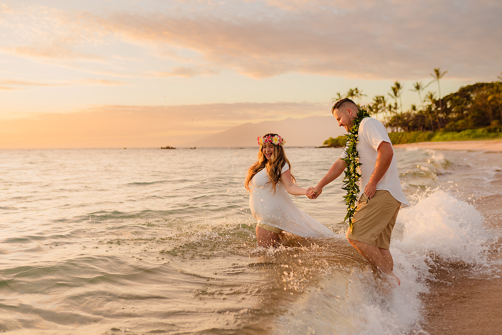 A couple holding hands while walking into the ocean and laughing, the woman is pregnant and wearing a long white dress and lei po'o and the man is wearing a lei during a maternity water photoshoot on maui. 