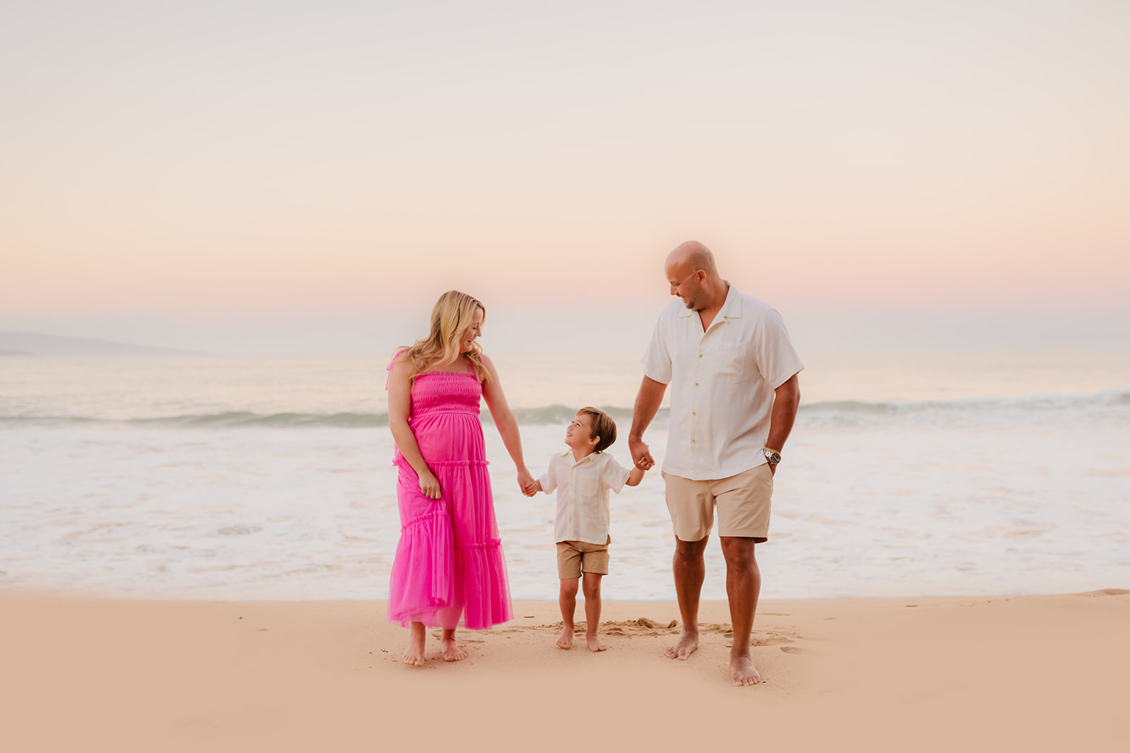 A woman and man hold their son's hands on the beach during a sunrise maternity family photoshoot on maui. 