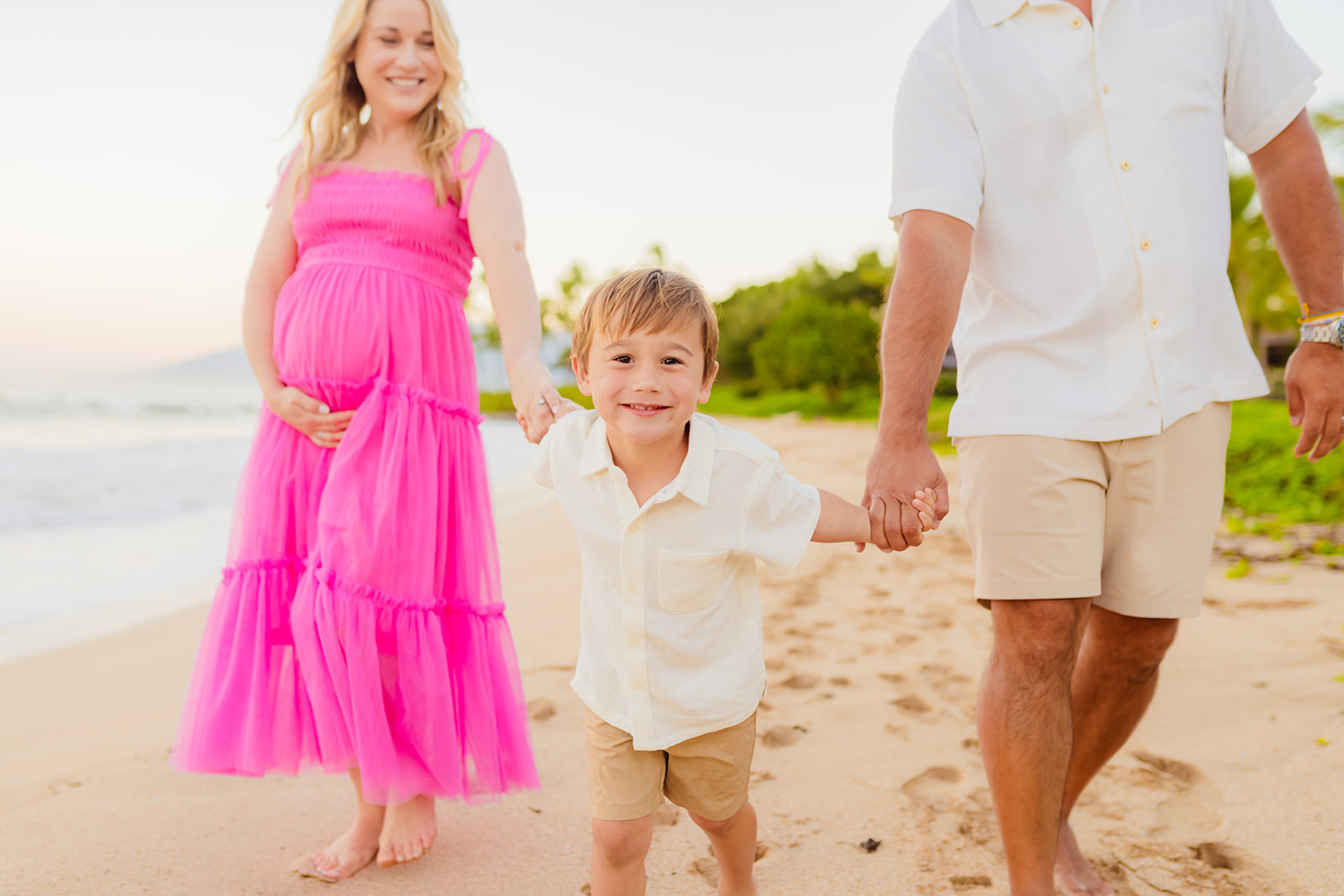 A young boy smiles while holding hands with his parents and walking the beach during a maui maternity photo session. 