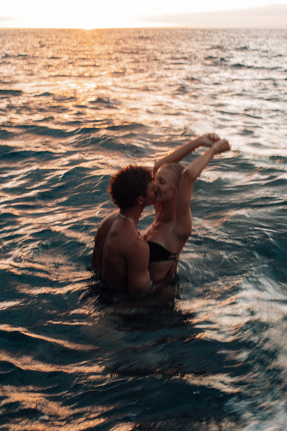 A couple embraces playfully in the gentle waves at sunset, the golden light casting a romantic glow over their joyful moment on a Maui beach. Photographed by Love and Water Photography.