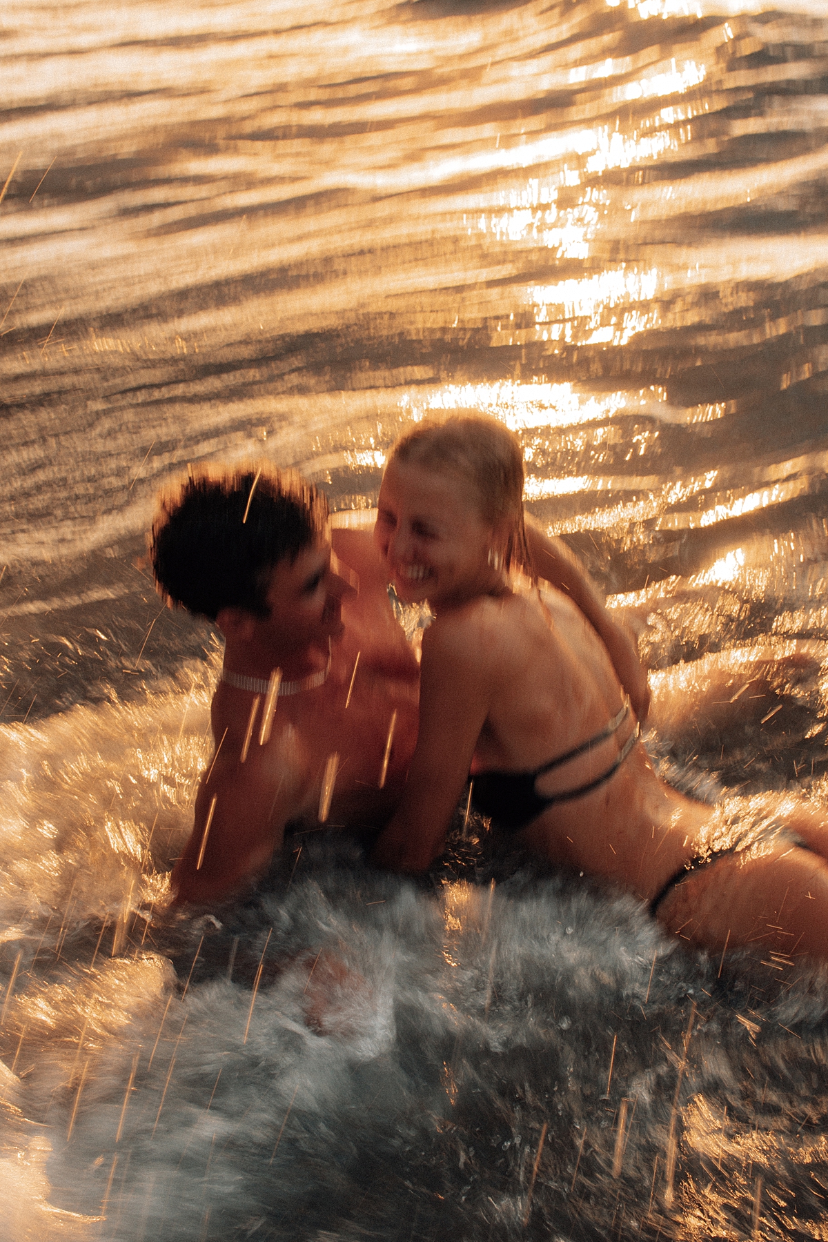 As the sun sets in Maui, a couple enjoys a playful moment in the waves, kissing and embracing as the water rushes around them. The scene is bathed in golden tones. Photographed by Love and Water Photography.