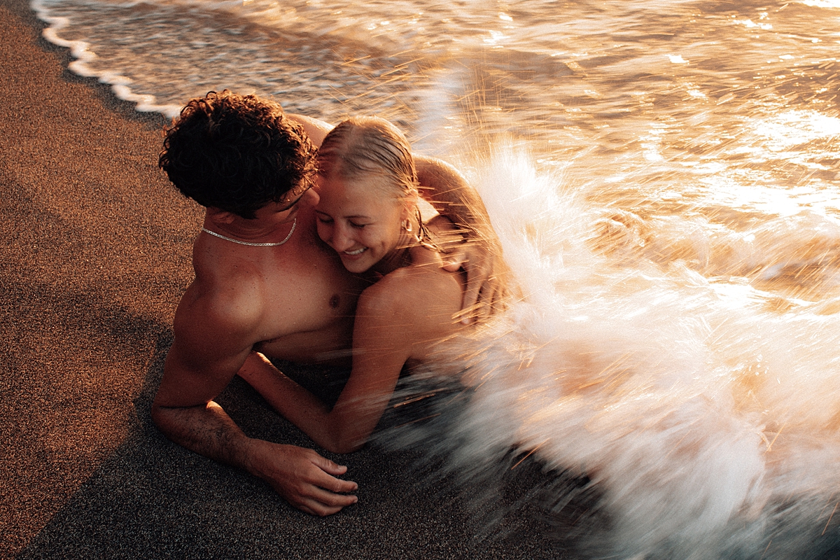 A playful couple shares a kiss in the Maui waves at sunset, surrounded by the warm glow of golden light as the water sparkles around them. Photographed by Love and Water Photography.