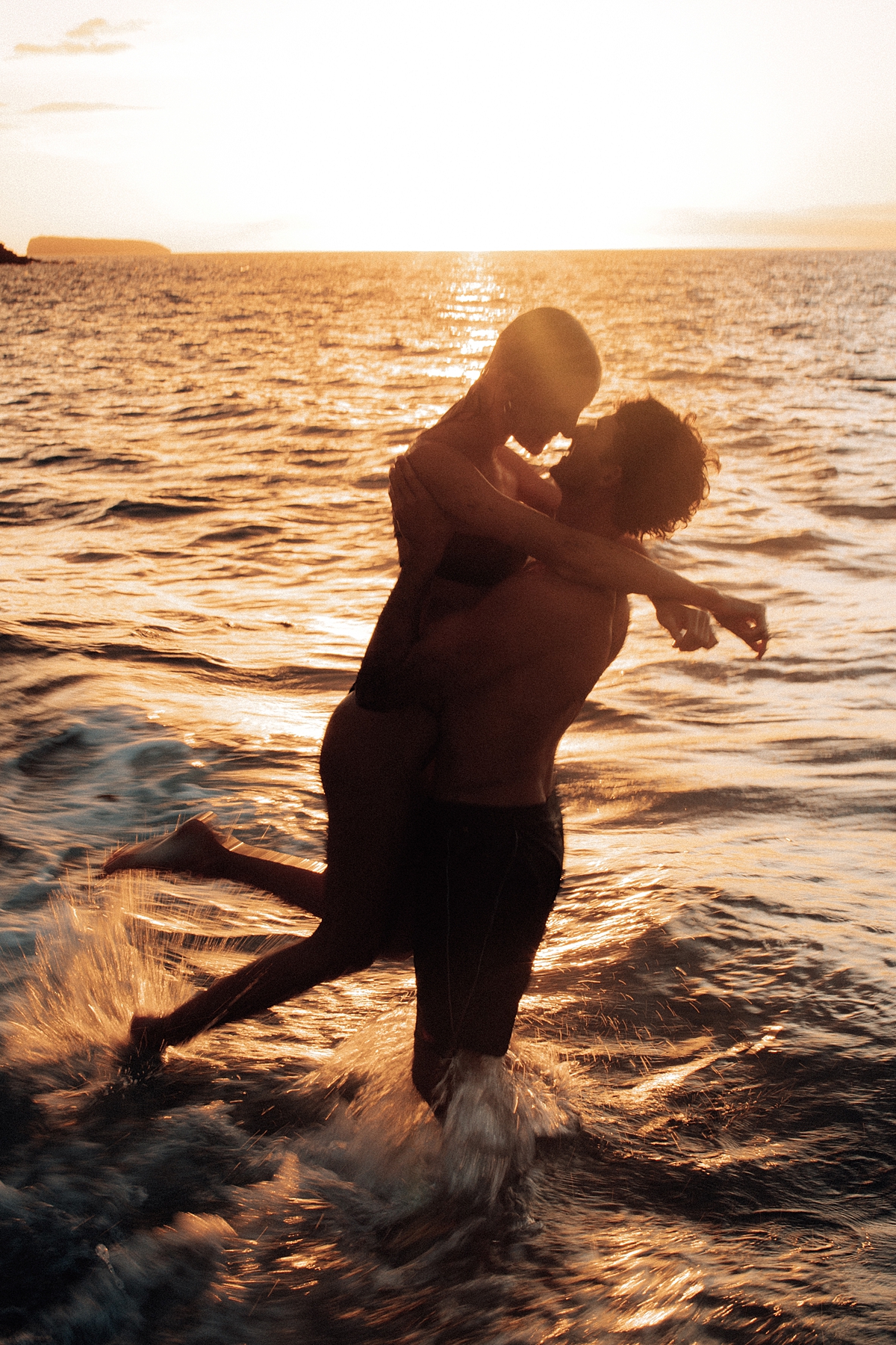 The warmth of the Maui sunset surrounds a couple as they kiss and play in the waves, their love radiating through the golden tones of the scene. Photographed by Love and Water Photography.