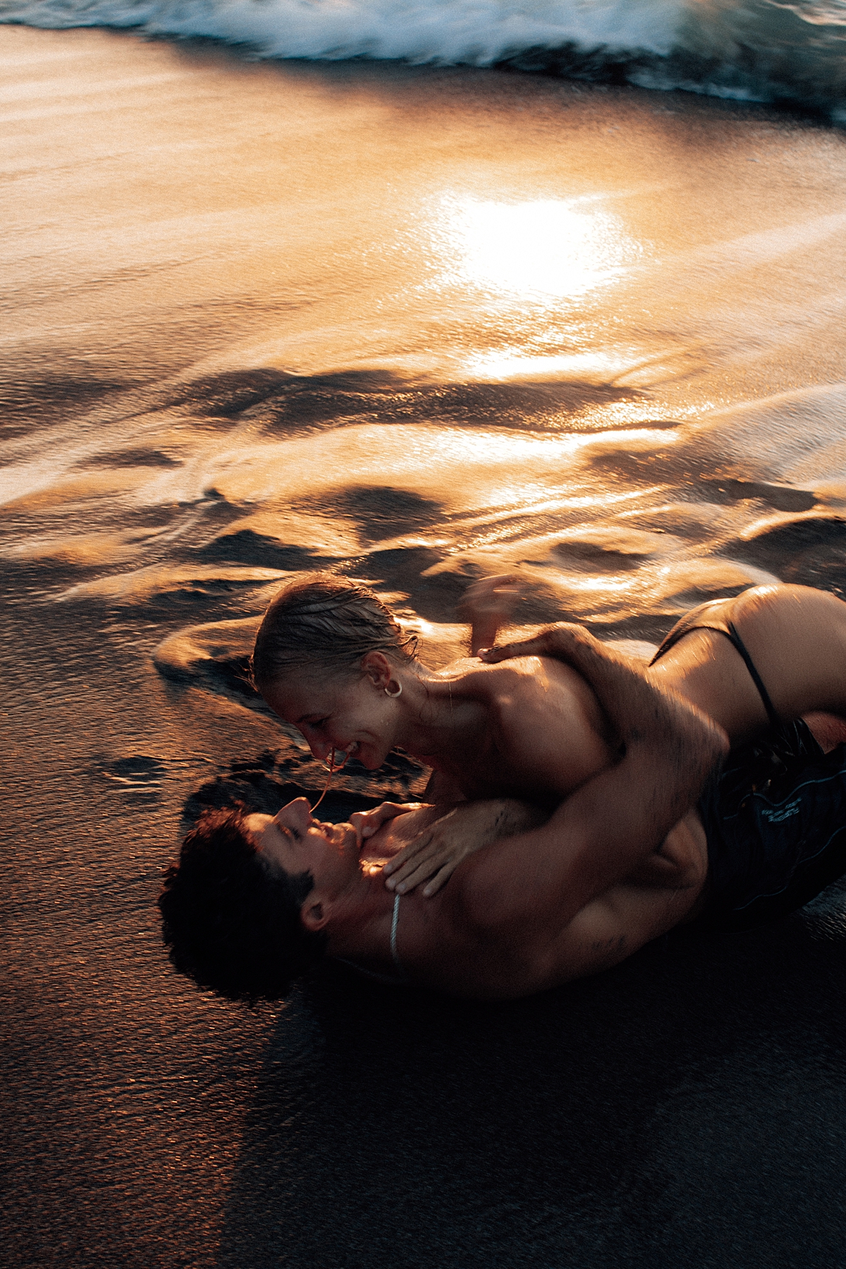 A couple shares a joyful moment in the ocean, kissing and holding each other as the Maui sunset fills the sky with golden light. The waves crash gently around them. Photographed by Love and Water Photography.