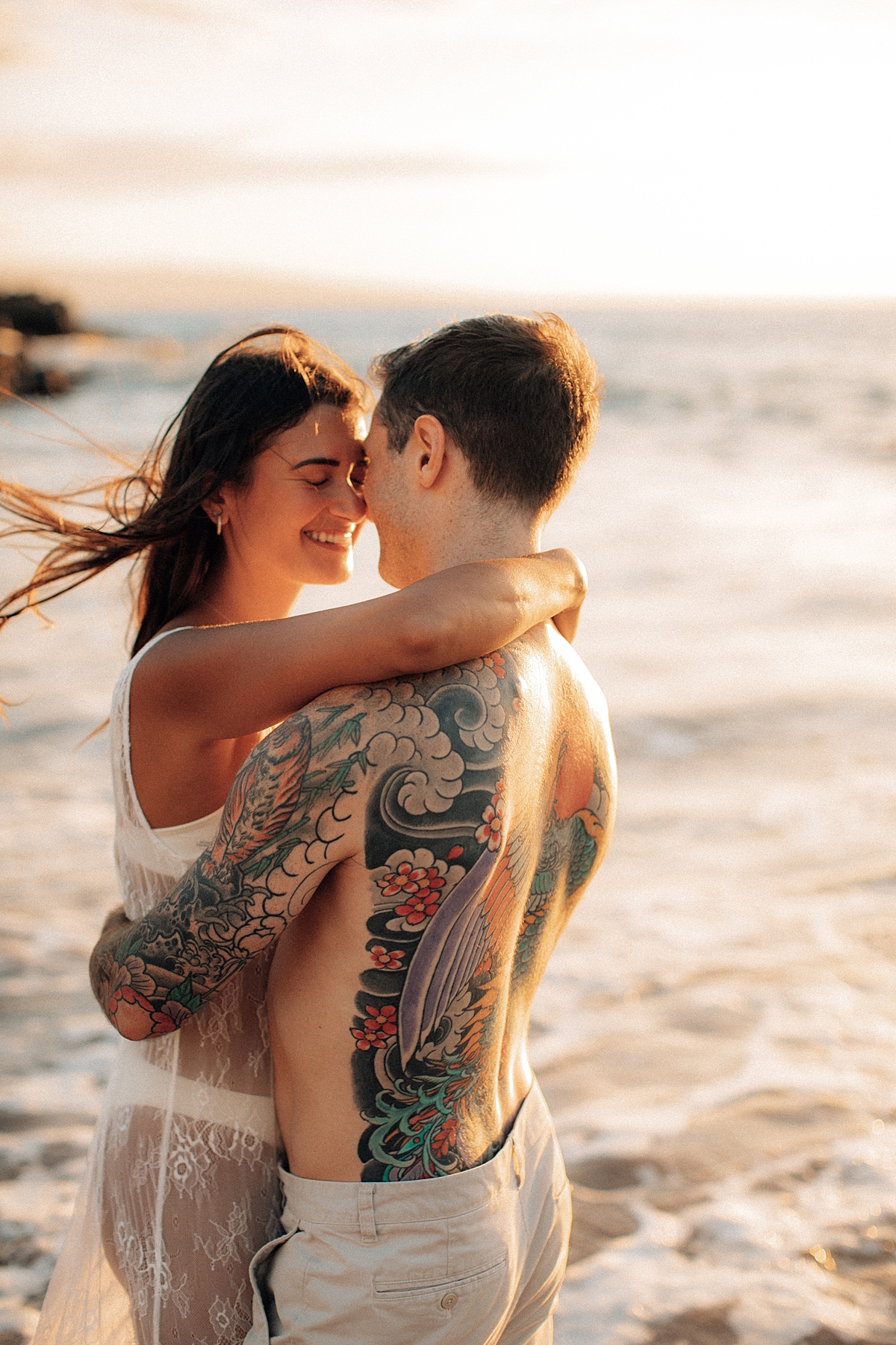 A woman smiles as her partner wraps his arms around her while they stand on the sand during a Maui beach photoshoot.