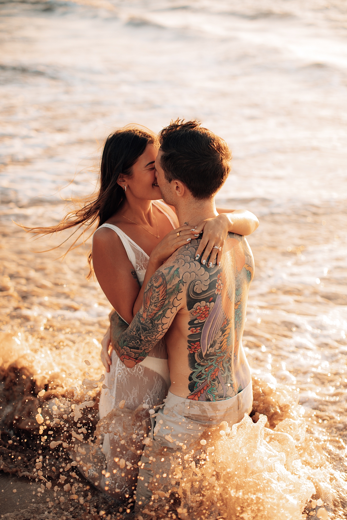 Playful couple embracing on the shoreline as they kneel in the sand, ocean waves nearby in Wailea, Maui.