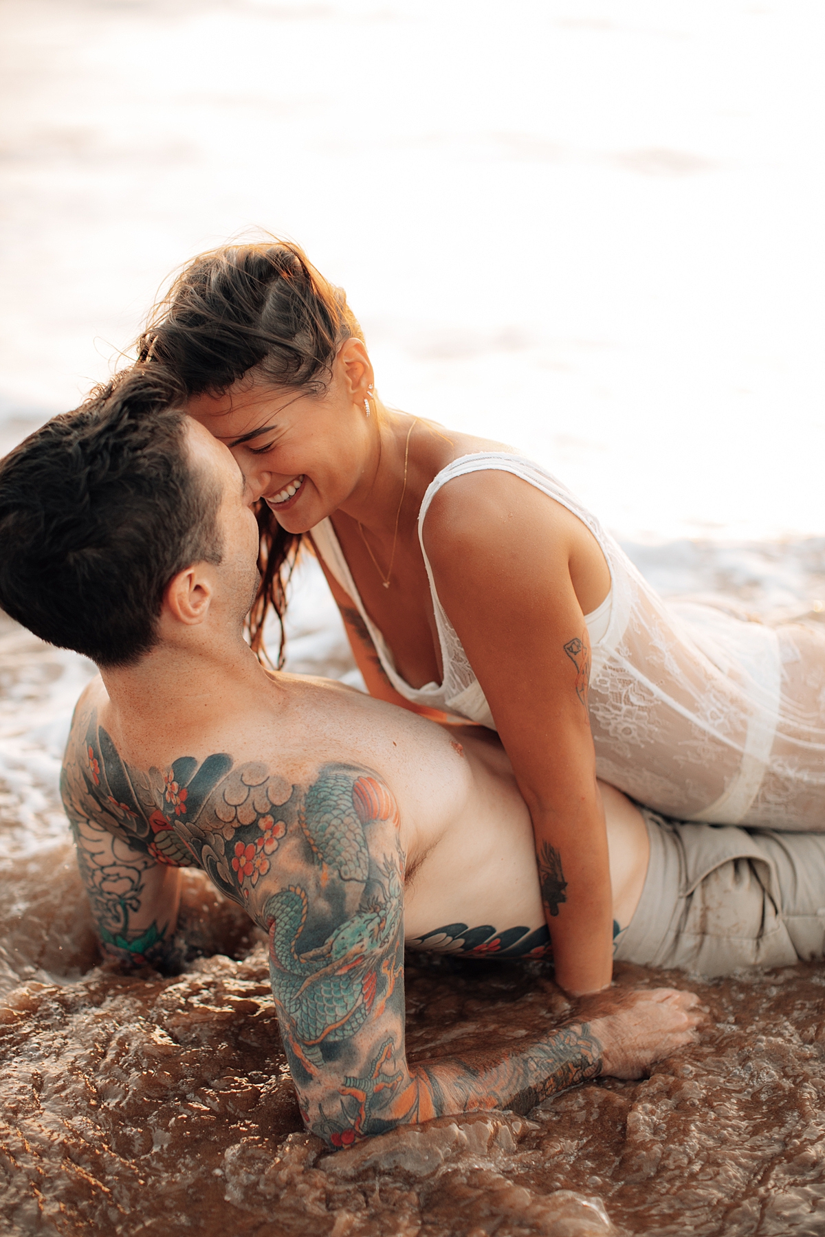 A man and woman laugh while rolling on the sandy beach at sunset in Wailea, Maui, in a candid moment by Love and Water.