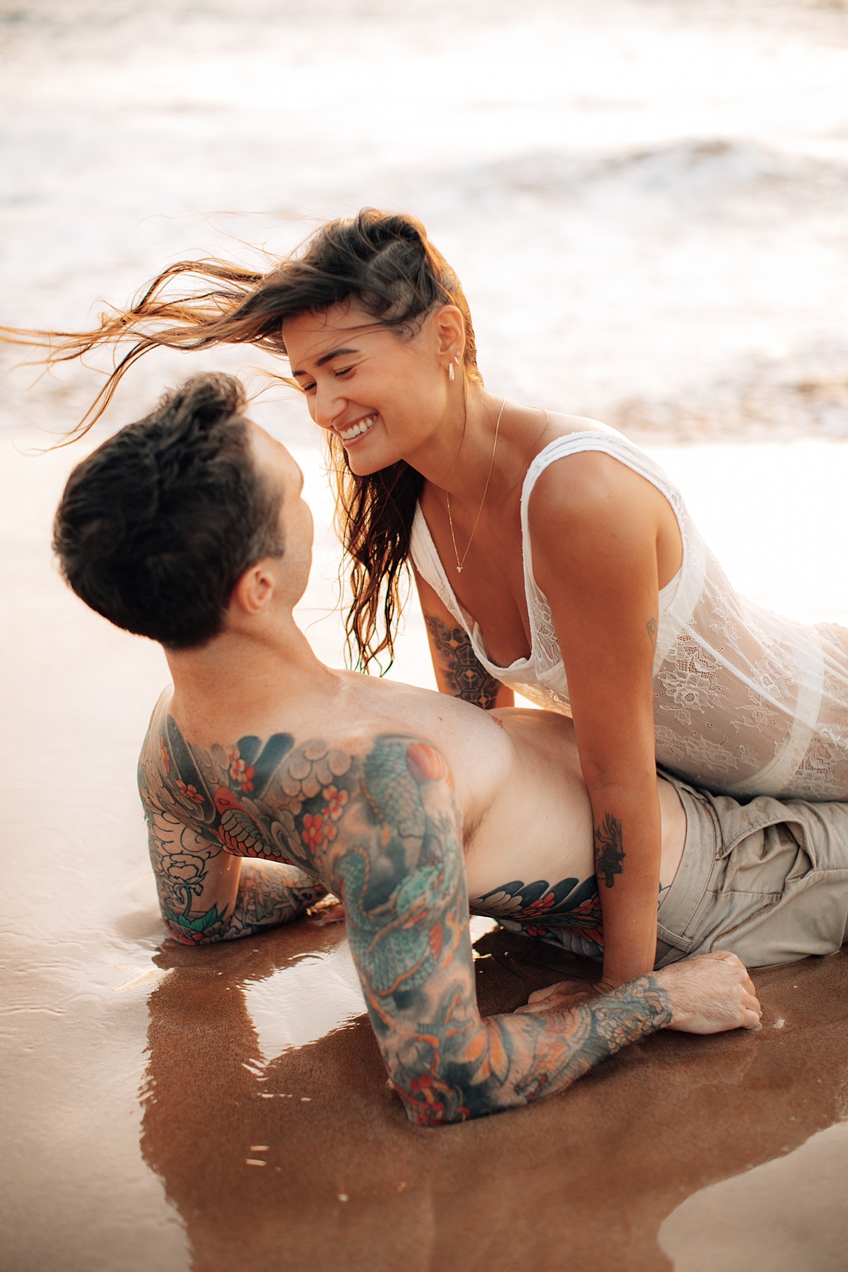 A couple joyfully tumbles on the sand with the turquoise ocean behind them in a Love and Water beach photoshoot.