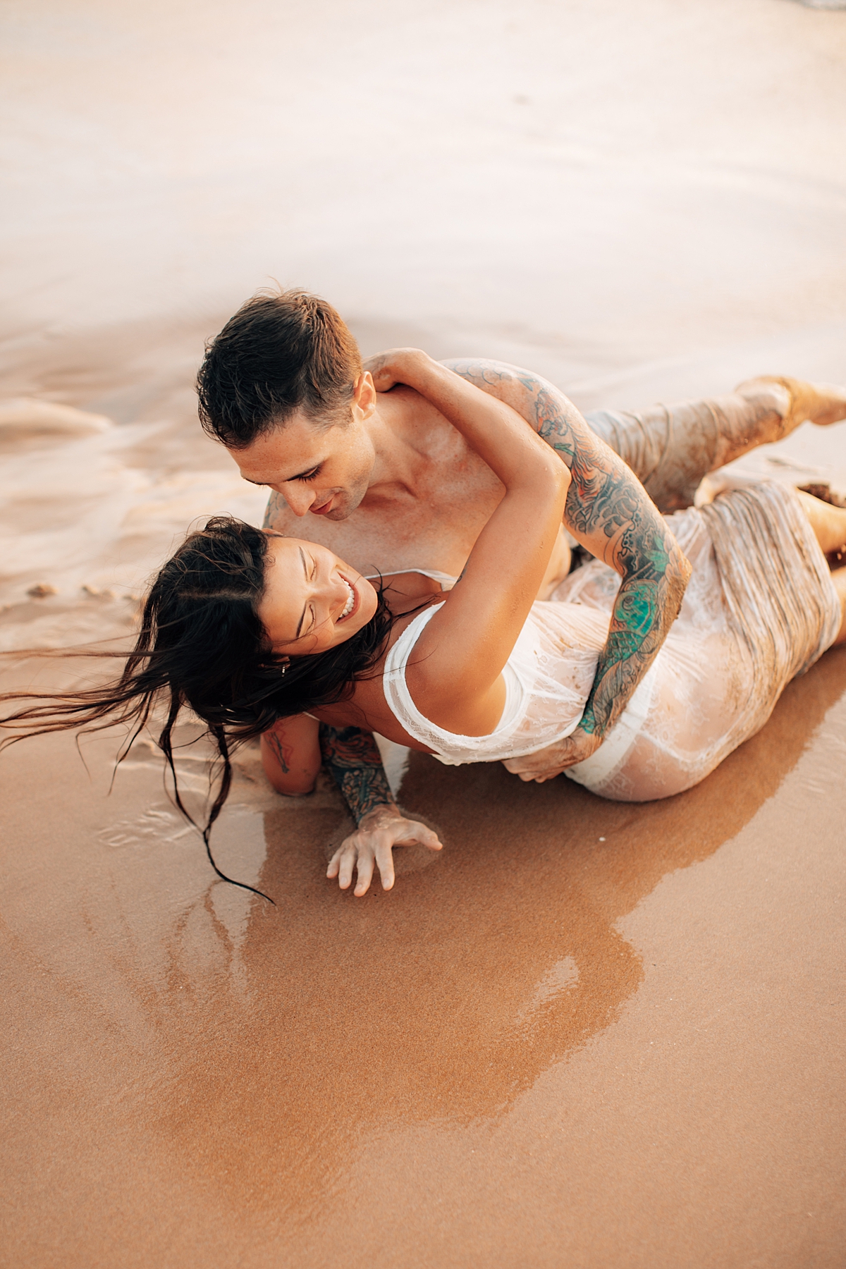 A couple lovingly embraces while lying on the sandy shore of a Maui beach, photographed by Love and Water.