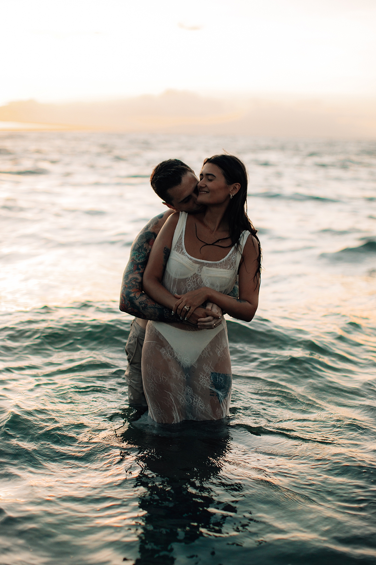 A couple, drenched in water, romantically embracing in the ocean, with the island coastline visible in the background.