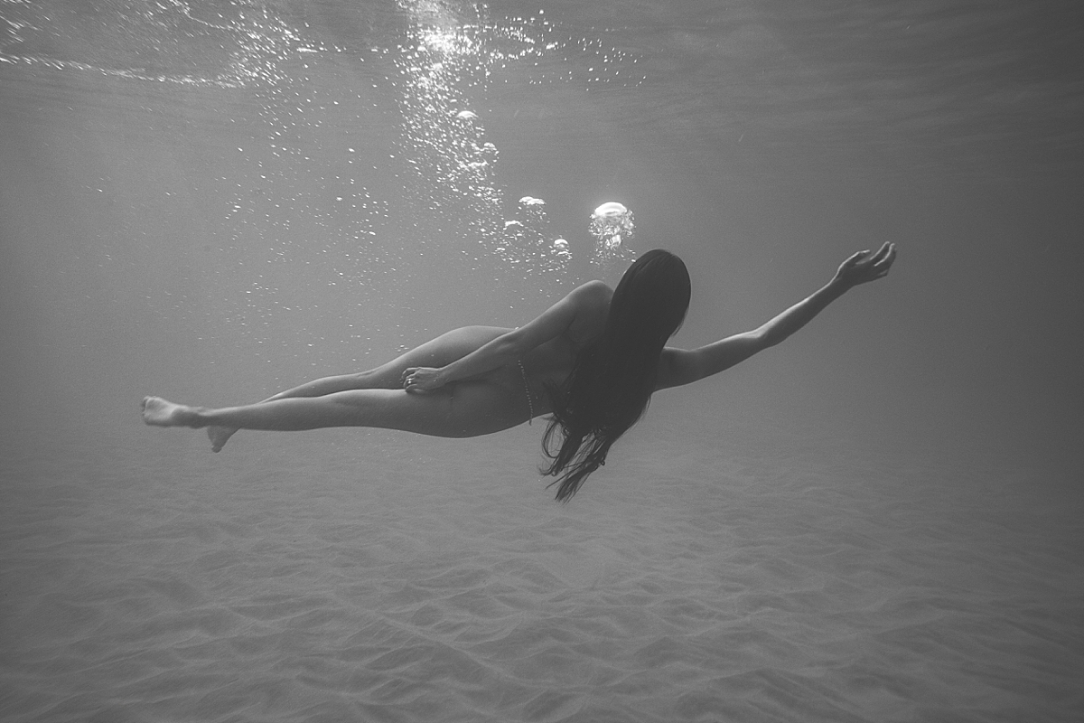 underwater black and white image of woman drifing in the sea with textured sand below her