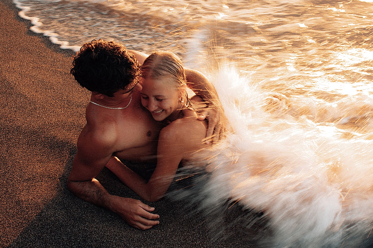 Waves splash a couple rolling in the ocean during their sunset photoshoot in Wailea