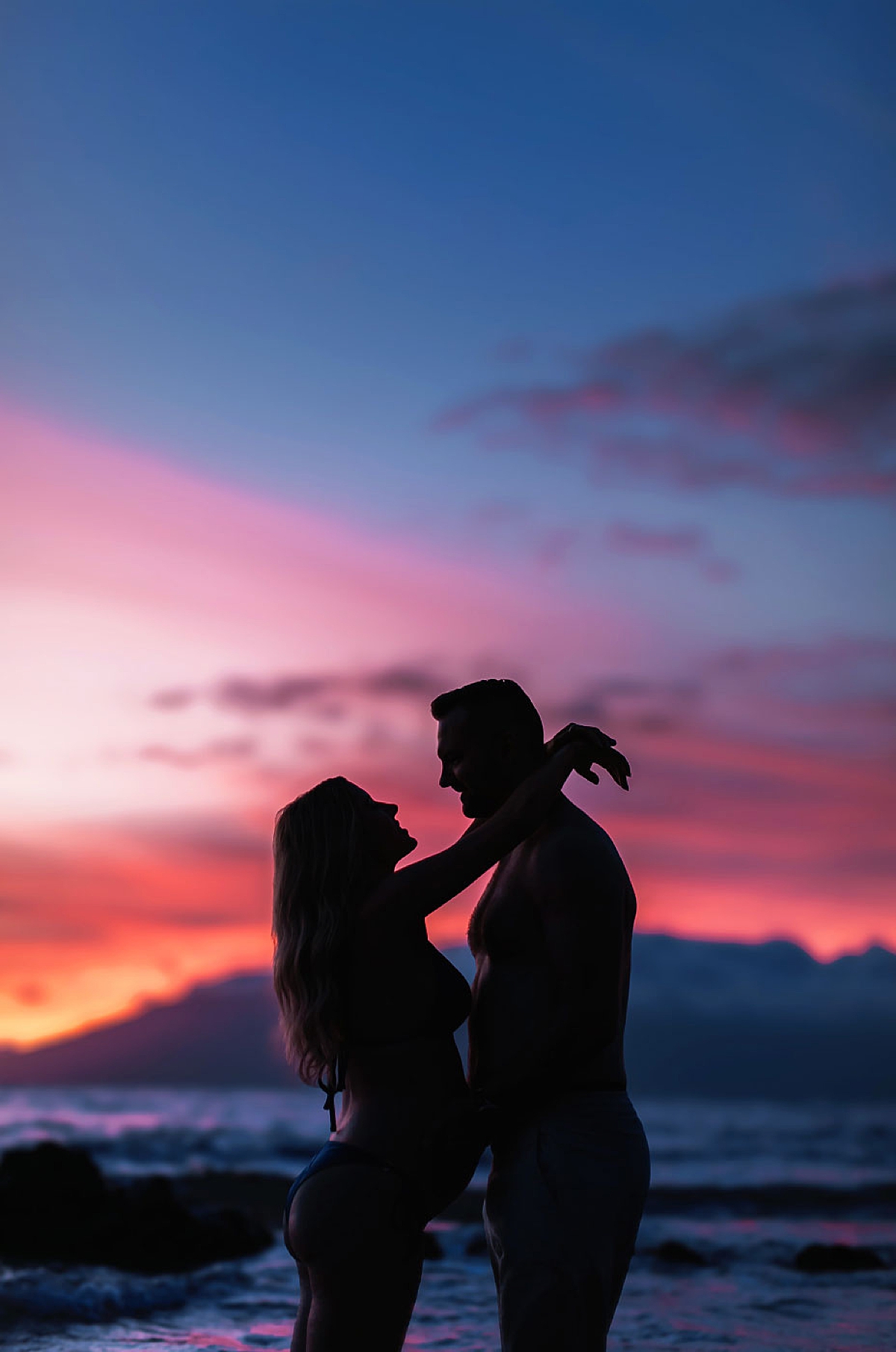 A couple shares a quiet moment by the shore as the sky turns a velvety pink and blue, framed by Maui’s calm evening ocean.