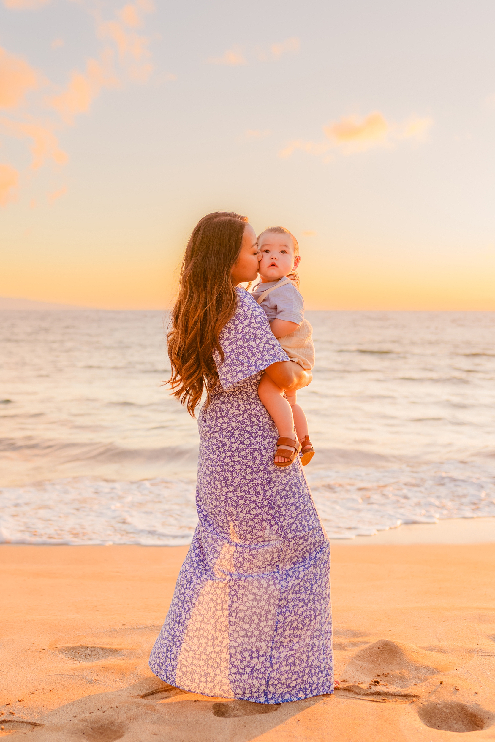 A mom and baby boy cuddling together on the Wailea shoreline as the Maui sunset lights up the background, captured by Love and Water Photography.