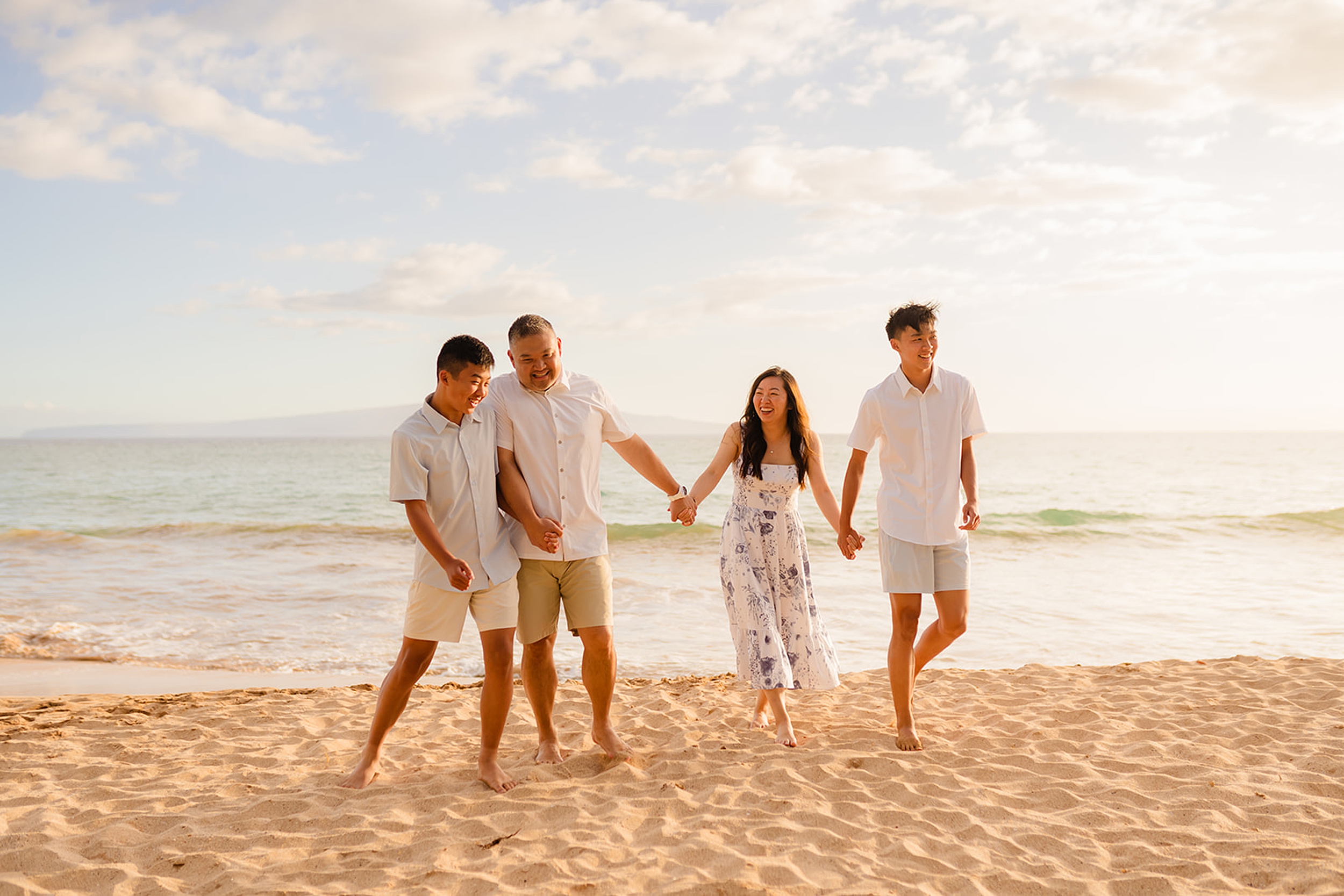 Family of four smiling together on the beach in Wailea during a sunset photoshoot, with gentle waves and warm light creating a serene backdrop.