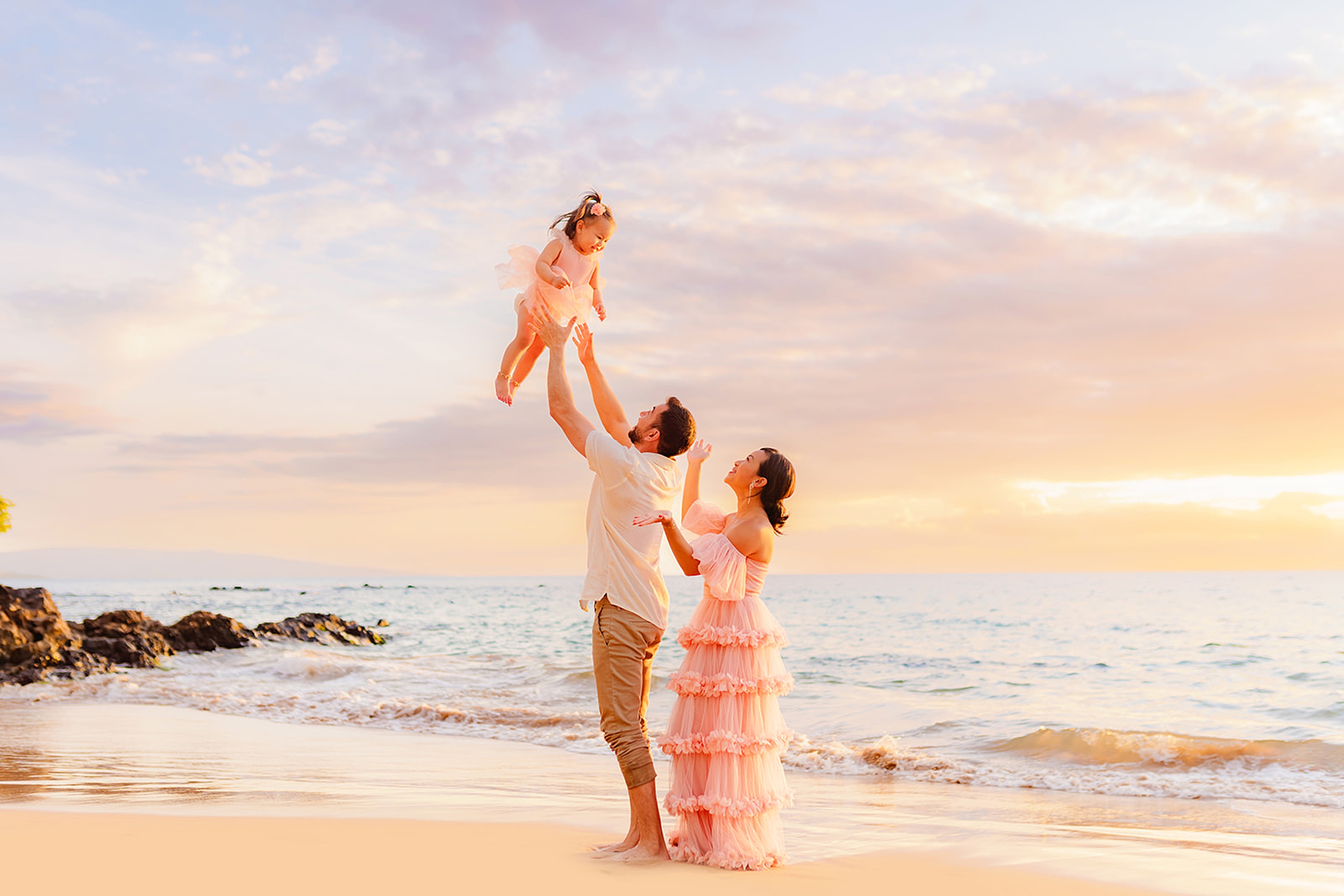 A toddler is tossed in the air by her dad in the sand with a scenic ocean view at White Rock Beach in Wailea, perfect for families staying at nearby Kihei condos.