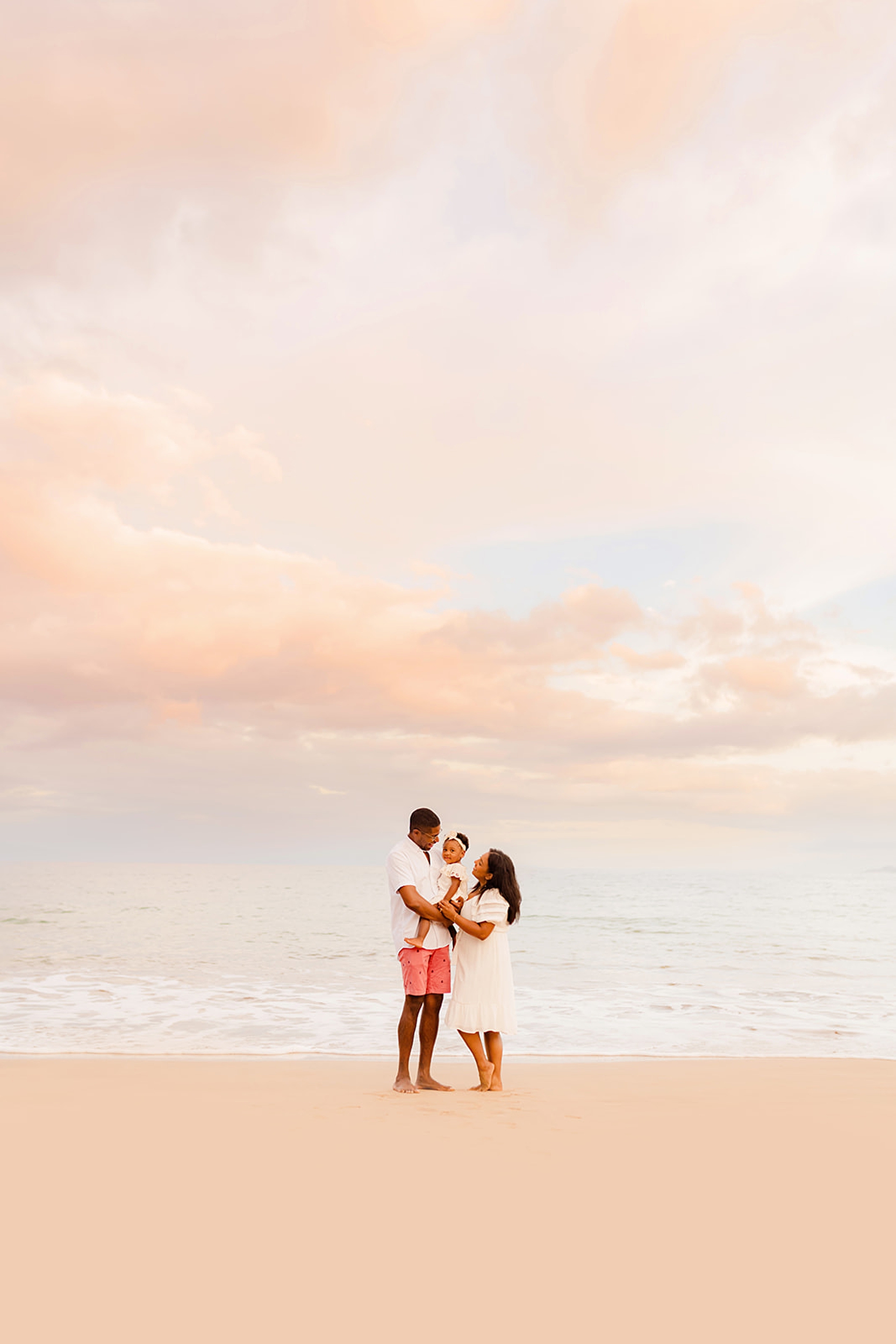 Parents holding their young baby girl close on the shoreline at the Four Seasons Wailea, captured by Love and Water Photography with the pastel Maui sunrise clouds behind them.