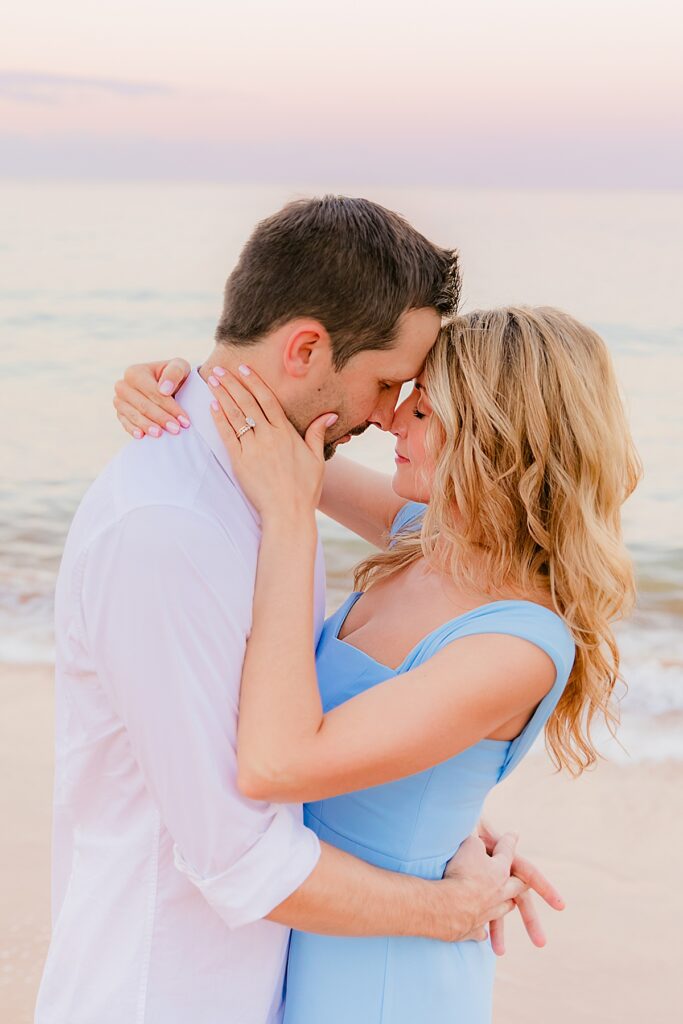 Woman and man with eyes closed holding one another close as a pastel sunrise unfolds behind them on the beach