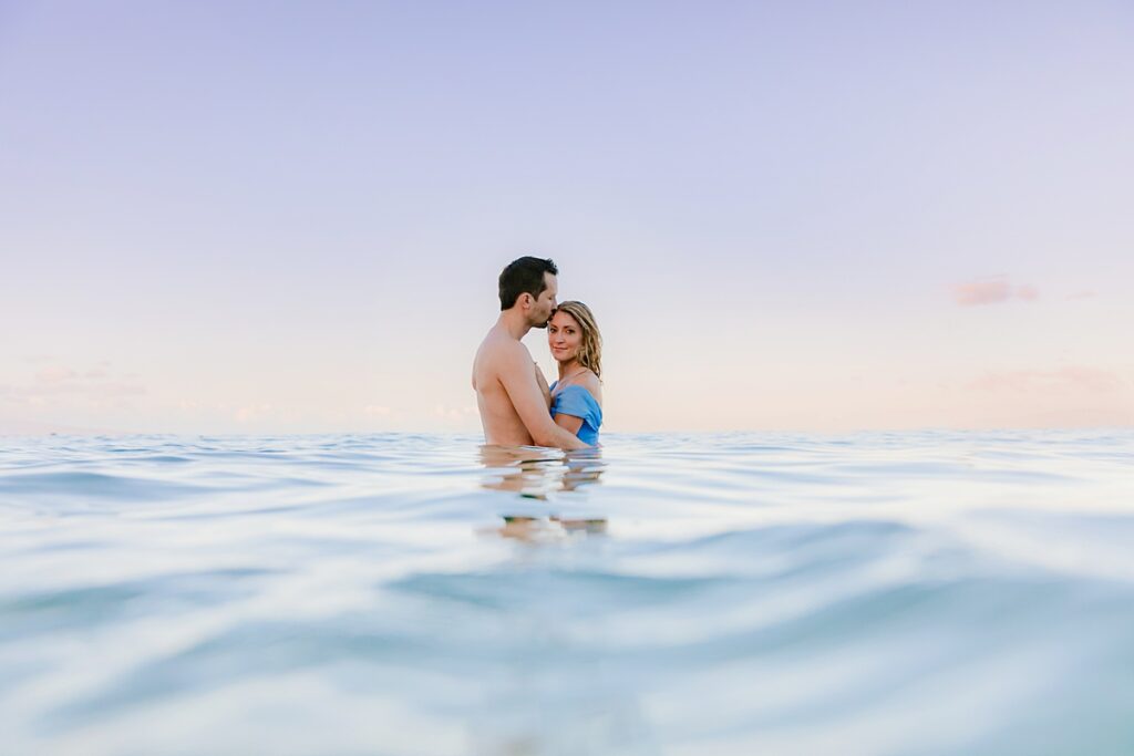 woman looks at the camera while standing in the ocean with her husband, who kisses her on the forehead