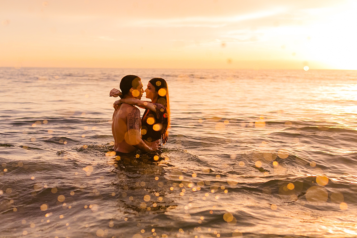 sunset creates bokeh as a couple splashes and embraces in the water at sunset.