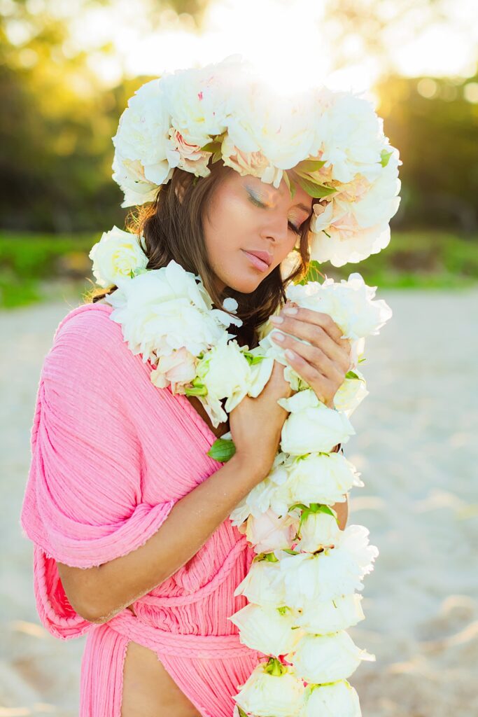 Close-up of a woman wearing a vibrant pink gown and a lei po'o adorned with fresh flowers, her face softly backlit by the golden hues of a Wailea sunrise.