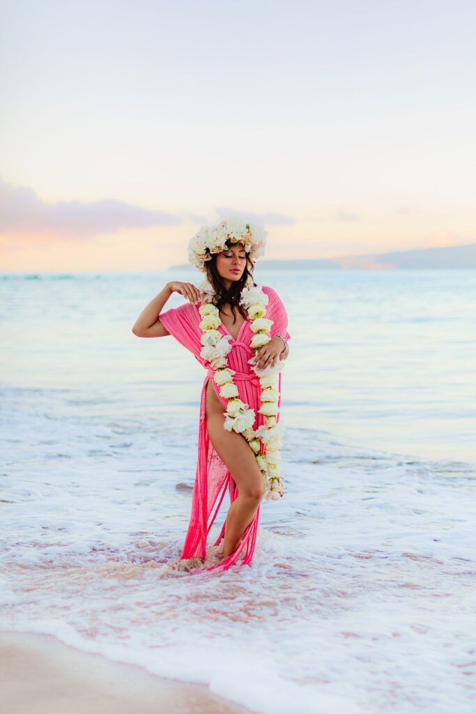 brown haired woman wearing a lei and pink gown, with the Maui sunrise creating a glowing backdrop of soft pastels during her beach portraits with love and water