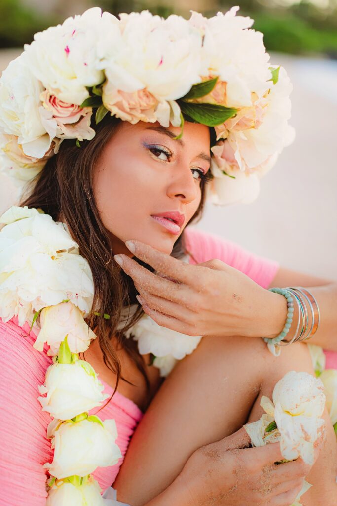 Woman sitting in the sand, her mermaid sparkle makeup catching the soft rays of sunrise, while her floral lei and lei po'o drapes elegantly over her shoulders and head.