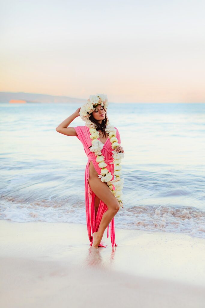 brunette woman in a flowing pink gown, her floral lei cascading down, as she closes her eyes at sunrise