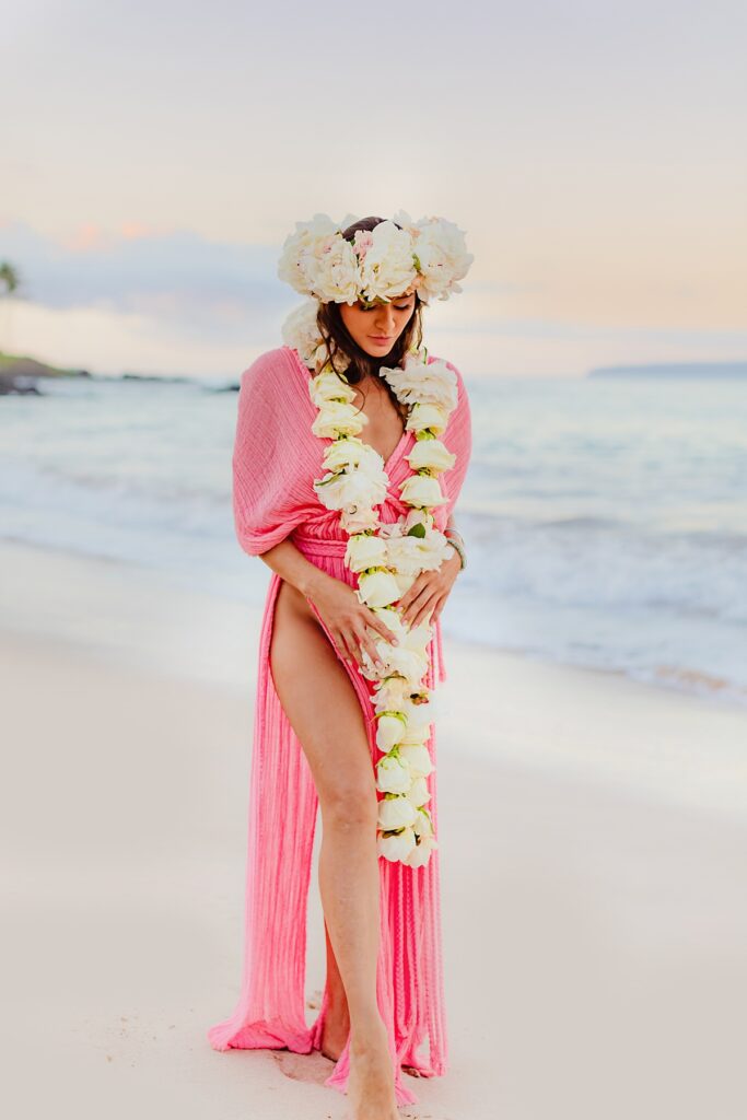 Woman standing and looking down in a pink Kasia Kulenty gown, her lei po’o catching the soft morning light on a serene beach in Wailea, Maui photographed by love and water